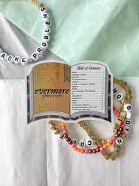 A decorative book-shaped pendant or sticker reading 'evermore (alternate version)' with an open-book design, styled on white and mint fabric with beaded necklaces and bracelets around it.