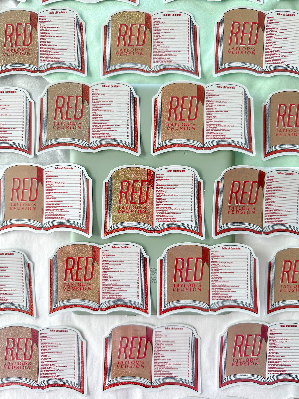 A repeating pattern of small book-shaped stickers or labels reading "RED" and "Taylor's Version" arranged on a pale mint background.