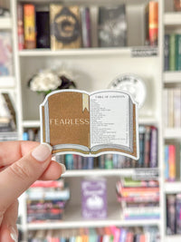 A hand holds a die-cut sticker shaped like an open book with a gold glittery cover and white interior page reading 'FEARLESS TABLE OF CONTENTS,' against a blurred bookshelf background.