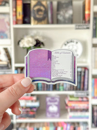 A hand holds a die-cut sticker shaped like an open book with a purple glittery cover and a white table of contents page, set against a blurred bookshelf background.