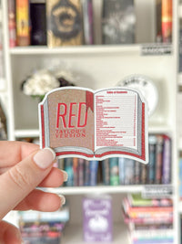 A hand holds a die-cut sticker shaped like an open book with the title 'RED TAYLOR VERSION' and a table of contents printed on it, set against a blurred bookshelf background.