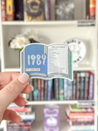 A hand holds a die-cut sticker shaped like an open book, featuring a blue glittery cover with '1987' and a white page with 'Table of Contents' text, against a blurred bookshelf background.
