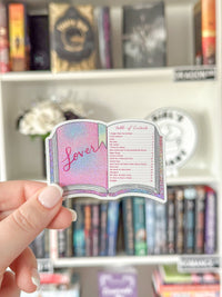 A hand holds a die-cut sticker shaped like an open book with a holographic pink and blue cover and the word 'Lover' written on it, set against a blurred bookshelf background.