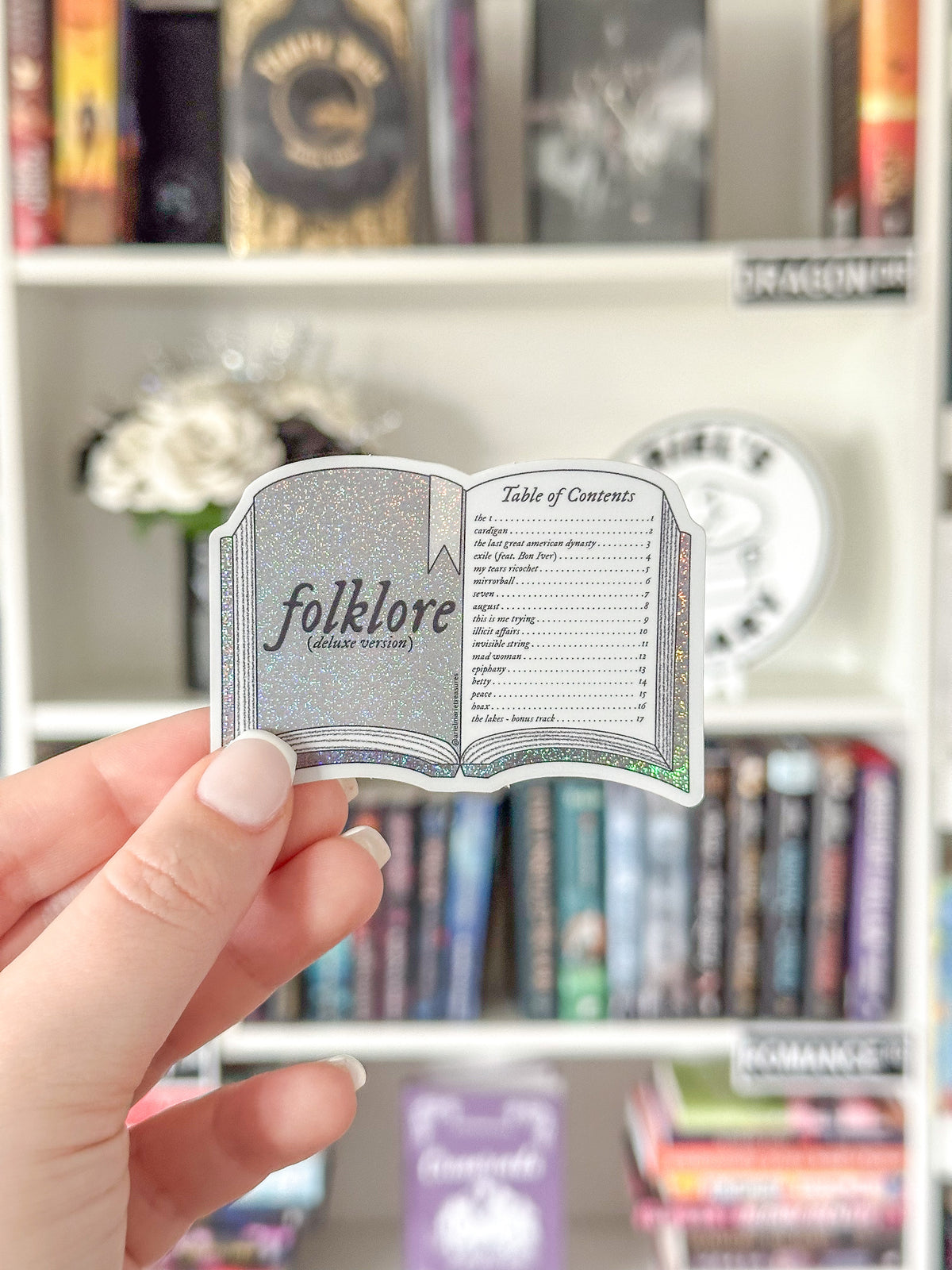 A hand holds a die-cut sticker shaped like an open book titled 'folklore (whate verison)' against a blurred background of a bookshelf.