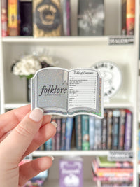 A hand holds a die-cut sticker shaped like an open book titled 'folklore (whate verison)' against a blurred background of a bookshelf.