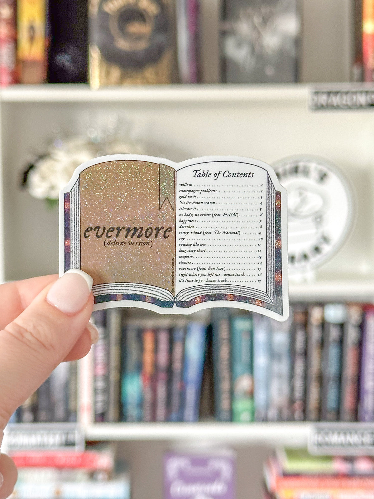 A hand holds a die-cut sticker shaped like an open book, featuring a glittery gold interior page with the text 'evermore (album cover)' and a printed table of contents design. The background shows blurred bookshelves with books and shelf labels.