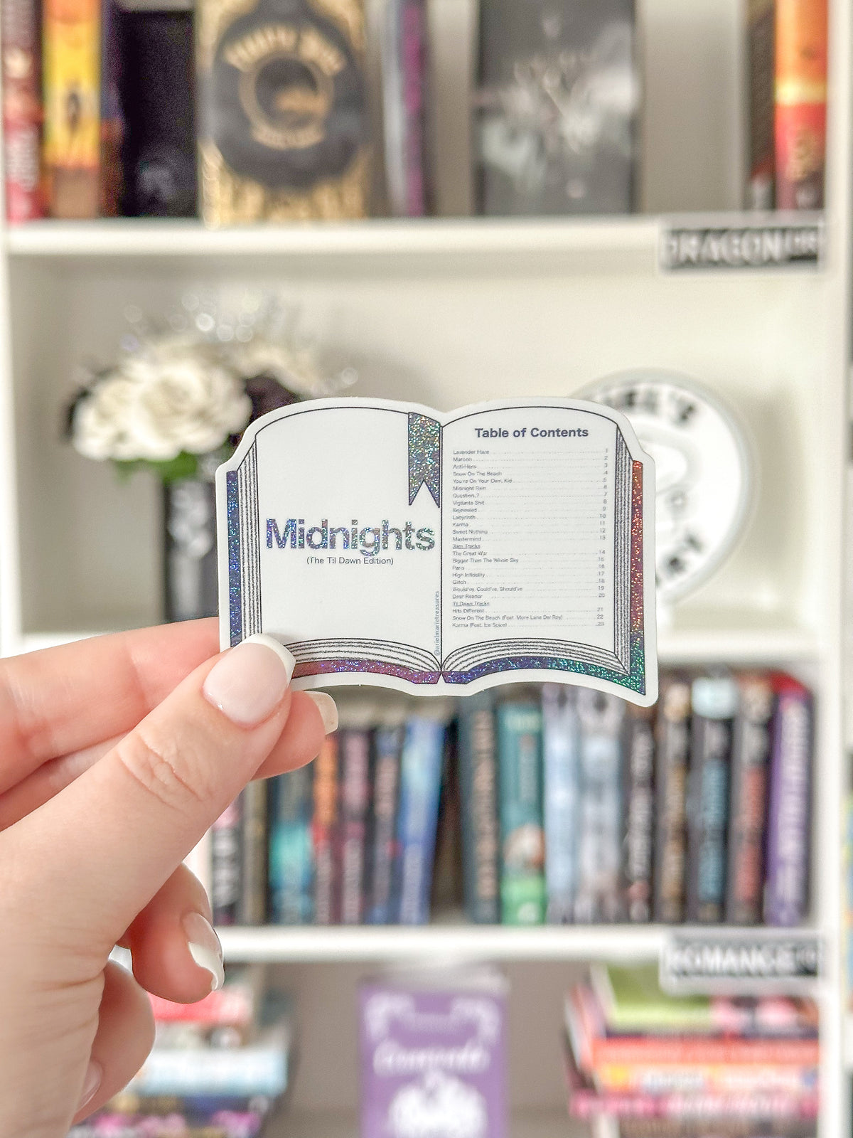 A hand holds a die-cut sticker shaped like an open book with the title 'Midnights' and a table of contents, set against a blurred bookshelf background.