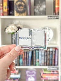 A hand holds a die-cut sticker shaped like an open book with the title 'Midnights' and a table of contents, set against a blurred bookshelf background.