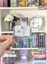 A hand with gray glitter nail polish holds a small die-cut sticker shaped like an open book, featuring the text 'The Tortuous Path's Department of Romance' and a monogram. The background shows a white bookshelf filled with romance novels and decorative items.