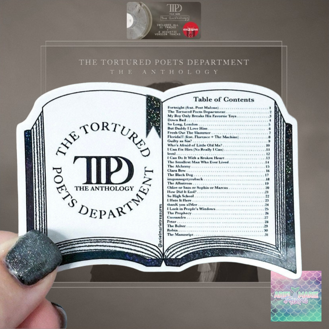 A hand with glittery dark nail polish holds a clear acrylic book-shaped plaque featuring the title 'The Tortured Poets Department: The Anthology' and an open-book design with printed text. A small round LP record and a pink-and-black album cover are visible above it against a taupe background.