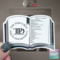 A hand with glittery dark nail polish holds a clear acrylic book-shaped plaque featuring the title 'The Tortured Poets Department: The Anthology' and an open-book design with printed text. A small round LP record and a pink-and-black album cover are visible above it against a taupe background.