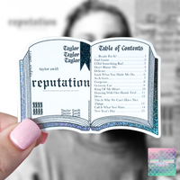 A hand with pink nails holds a die-cut sticker shaped like an open book titled 'reputation' with Taylor Swift and Taylor Taylor text, against a blurred gray background. A small logo in the bottom right reads 'Taylor Swift Fan Club'.