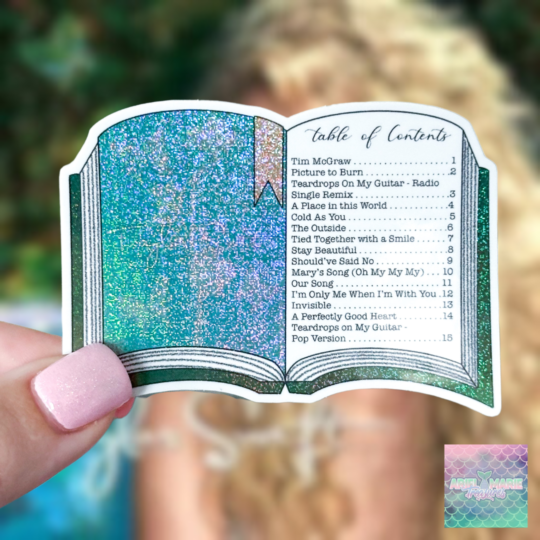 A hand with pink glitter nail polish holds a die-cut sticker shaped like an open book. The sticker features a glittery teal and blue interior with a pink bookmark and printed table of contents text.