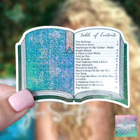 A hand with pink glitter nail polish holds a die-cut sticker shaped like an open book. The sticker features a glittery teal and blue interior with a pink bookmark and printed table of contents text.