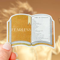 A die-cut sticker shaped like an open book with a glittery gold cover and a white table of contents page, held by a hand against a warm beige background.