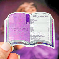 A die-cut sticker shaped like an open book cover, featuring a glittery purple cover with the words 'Speak Now Taylor's Version' and a table of contents on the right page. The sticker is held by a hand against a blurred purple background.