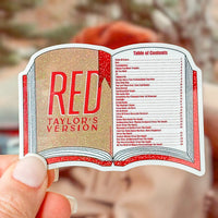 A hand holding a die-cut sticker shaped like an open book, featuring the title 'RED TAYLOR'S VERSION' in large red letters on a glittery gold cover page with a table of contents printed on the second page. The background is a soft, blurred outdoor scene.
