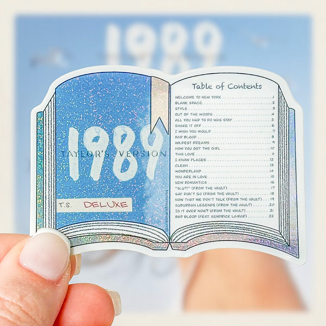 A hand holding a die-cut sticker shaped like an open book, featuring a glittery blue cover with '1989' and a table of contents page. The sticker has a holographic/silver border and reads 'T.