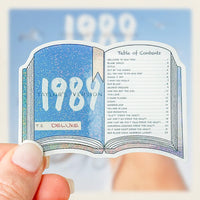 A hand holding a die-cut sticker shaped like an open book, featuring a glittery blue cover with '1989' and a table of contents page. The sticker has a holographic/silver border and reads 'T.