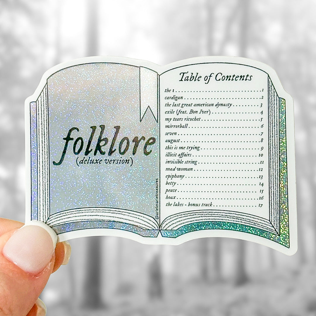 A die-cut sticker shaped like an open book with the title 'folklore (deluxe version)' on the left page and a 'Table of Contents' on the right page, held between fingers against a blurred outdoor background.