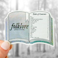 A die-cut sticker shaped like an open book with the title 'folklore (deluxe version)' on the left page and a 'Table of Contents' on the right page, held between fingers against a blurred outdoor background.