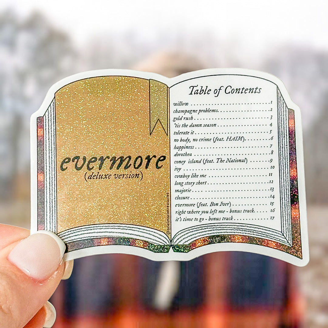 A hand holds a die-cut sticker shaped like an open book, featuring glittery gold and beige cover pages with black text reading 'evermore (deluxe version)' and a printed table of contents on the right page. The background is a softly blurred outdoor scene.