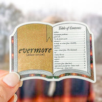 A hand holds a die-cut sticker shaped like an open book, featuring glittery gold and beige cover pages with black text reading 'evermore (deluxe version)' and a printed table of contents on the right page. The background is a softly blurred outdoor scene.