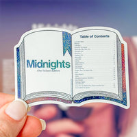 A hand holding a die-cut sticker shaped like an open book showing a table of contents page for 'Midnights (The Til Dawn Edition)'. The sticker features a white background with blue glitter accents on the ribbon bookmark and page borders, and pink glitter accents on the right margin.