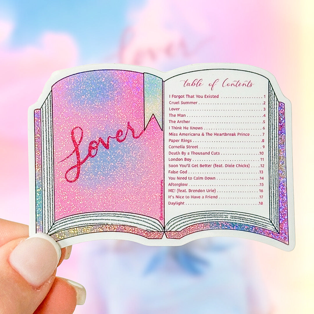 A hand holds a die-cut sticker shaped like an open book with glittery pink and purple pages, featuring the word 'Lover' on the left page and a table of contents on the right. The background is a soft pastel sky gradient.