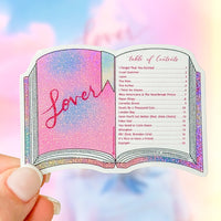 A hand holds a die-cut sticker shaped like an open book with glittery pink and purple pages, featuring the word 'Lover' on the left page and a table of contents on the right. The background is a soft pastel sky gradient.